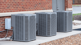 Three outdoor air conditioning units on a concrete pad beside a brick building, with wiring and gravel nearby.