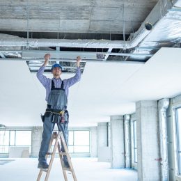 commercial hvac technician installing ductwork above a white drop ceiling