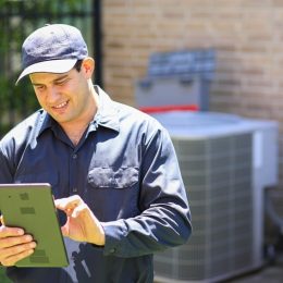 HVAC technician in the foreground looking at a tablet with an a/c unit in the background