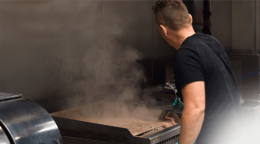 Man standing at a barbecue grill, tending meat as steam and smoke rise from the sizzling food.