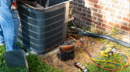 Person repairing an outdoor air conditioning unit with tools and electrical cables on the ground.