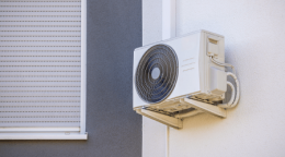 Outdoor air conditioner condenser unit mounted on a white exterior wall beside a window with a closed shutter.
