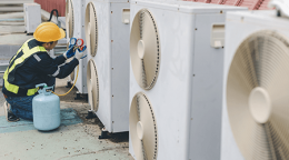 HVAC technician in safety gear servicing outdoor air conditioning units, with a blue refrigerant cylinder nearby.