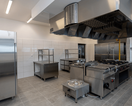 Large commercial kitchen with stainless steel stoves, exhaust hood, and prep tables against tiled walls.
