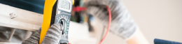 Person wearing gloves using a digital multimeter to test electrical circuits, yellow device held near a panel.