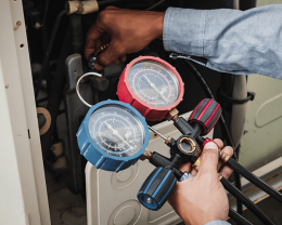 HVAC technician’s hands adjusting a red and blue manifold gauge set connected to refrigerant lines to measure pressure at an air conditioner equipment.