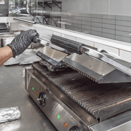Gloved hand holding a metal grill press on a commercial stainless steel griddle to press and cook food from above.