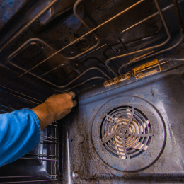 Person in a blue sleeve reaches into the dirty oven, touching a rack inside the metal cavity with coils and a circular fan at the bottom