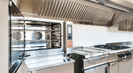 Industrial commercial kitchen with stainless steel appliances, including a double stacked oven with a digital control panel and an overhead range hood above a gas stove and flat griddle.