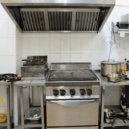 Commercial kitchen with stainless steel stove, flat-top griddle, fryer, and overhead exhaust hood; pots and pans on the counters.