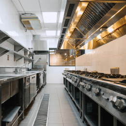 Commercial kitchen with stainless steel appliances on both sides, a long row of gas stoves on the right and prep counters on the left under overhead ventilation hood.