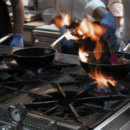 Chefs in a stainless kitchen cook on a large gas stove, flames leaping from two frying pans on the burner of a professional setup.