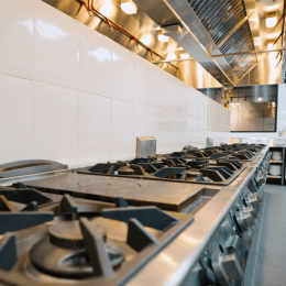 Commercial stainless steel gas range with multiple burners in a professional kitchen, under bright overhead vents and white tiled wall.