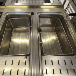 Two side-by-side commercial deep fryers in a stainless steel kitchen, with metal baskets and black handles resting inside each fryer.