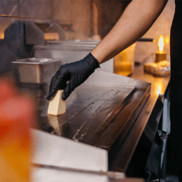 Chef wearing a black glove places a slice of cheese on a hot griddle in a busy kitchen.