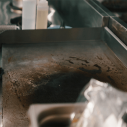 A dirty flat-top griddle with oil stains and burnt residue in a stainless kitchen, suggesting a prep area after cooking.