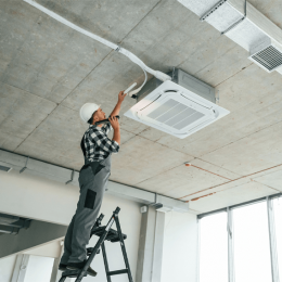 HVAC technician on a ladder installing a ceiling-mounted air conditioner unit in a concrete-open space
