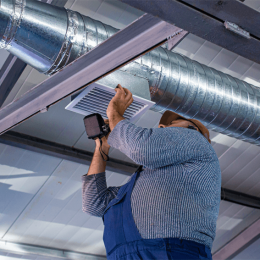 HVAC technician in blue overalls installing a square ceiling air vent beneath exposed metal ductwork in an industrial setting