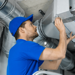 HVAC technician in a blue uniform inspecting an air duct in a mechanical room