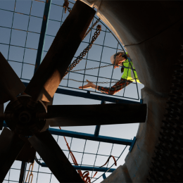 Worker in a high-visibility vest climbs inside a large circular industrial duct, supported by chains, with metal blades and blue framework visible.