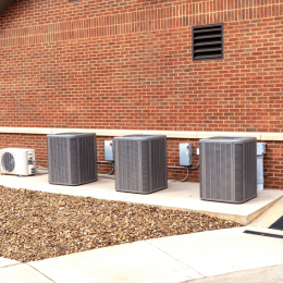 Three large outdoor HVAC condenser units lined up along a brick wall on a concrete pad, with gravel in the foreground.