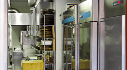 Commercial kitchen with stainless steel ovens, racks of trays, and prep area in the background.