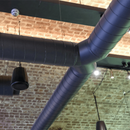 Industrial ceiling with exposed brick wall, two large blue HVAC ducts crossing overhead, and hanging pendant lights.