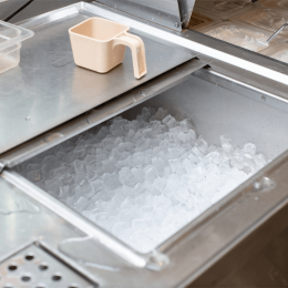 Ice bin filled with clear ice cubes in a stainless steel trough; a beige plastic pitcher sits on the lid nearby.
