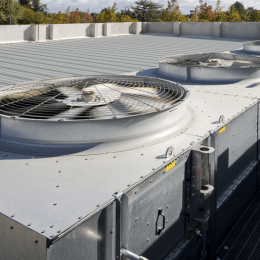 Rooftop HVAC unit with two large circular metal fans behind protective grilles on a flat metal roof; trees visible in the background.