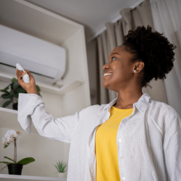Smiling woman with natural hair points a remote at a wall-mounted air conditioner in a bright, plant-filled room.