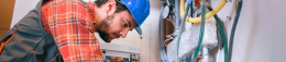 Tradesperson in a blue hard hat and red plaid shirt inspecting a control panel among pipes and wiring in a workshop.