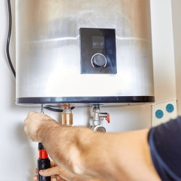 Technician tightening fittings on a wall-mounted stainless steel water heater with a screwdriver.