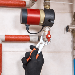 Gloved hand with orange-handled pliers turning a small red valve on a metal pipe with orange insulation.