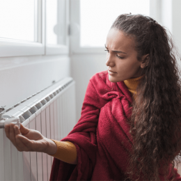 Woman in a red robe touches a radiator, looking concerned about the cold.