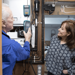 A male technician in a blue shirt uses a diagnostic meter on a boiler while a smiling woman stands nearby in a patterned blouse.