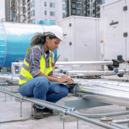 Female worker in a hard hat and high-visibility vest crouches on an industrial rooftop, typing on a laptop next to metal pipes and equipment with an urban backdrop.