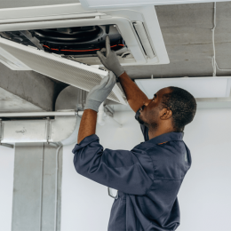 Maintenance worker in a navy jumpsuit and gloves services a ceiling-mounted HVAC unit in a commercial space.
