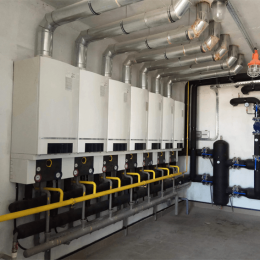 Row of white industrial boilers mounted along a wall with yellow gas pipes and large overhead ducts in a utility room.