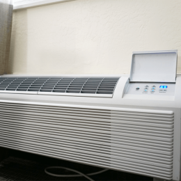 Window air conditioner unit installed in a wall with vents across the front and a control panel on the right side showing illuminated LEDs.