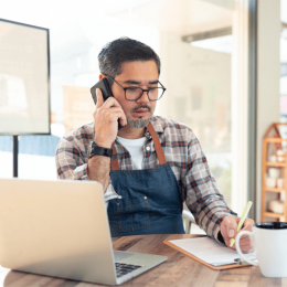 Man in plaid shirt and blue apron on a call at a desk with a laptop, writing on a notepad.