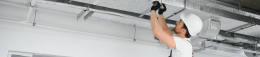 Male construction worker in a white hard hat and gloves tightening ductwork on an exposed ceiling HVAC system