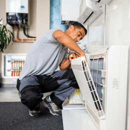 Technician crouching in a modern kitchen, repairing a white front-loading washing machine with its panel open and tools in hand.