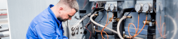 Maintenance technician in blue coveralls inspecting an industrial control panel with gauges and hoses