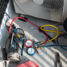 HVAC refrigerant manifold with red and blue hoses attached to gauges, on a metal floor near an outdoor condenser unit.