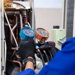 Technician's gloved hands hold two pressure gauges connected to hoses, testing an HVAC system.